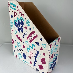 May include: Cardboard magazine holder with a cream-colored exterior. The holder is decorated with a repeating pattern of geometric shapes in teal, purple, and navy blue. The interior is plain brown.