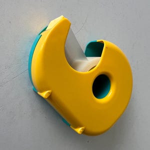 May include: A yellow and teal tape dispenser. The dispenser has a rounded shape with a teal accent on the sides and a circular hole. The tape roll is visible inside the dispenser.