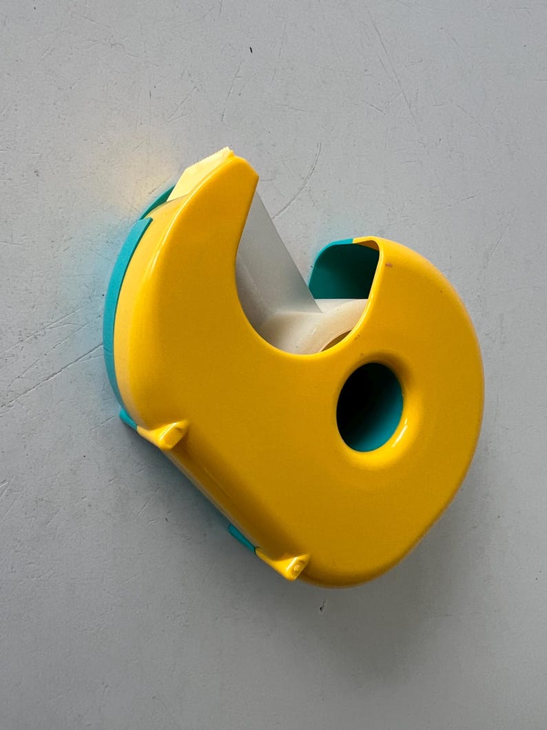 May include: A yellow and teal tape dispenser. The dispenser has a rounded shape with a teal accent on the sides and a circular hole. The tape roll is visible inside the dispenser.