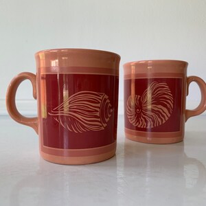 May include: Two ceramic mugs with a pink glaze and red accents. One mug features a seashell design, while the other has a nautilus shell design.