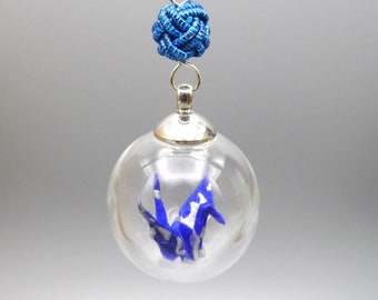 Può includere: Una collana con catena d'argento con una gru origami blu all'interno di un ciondolo di vetro trasparente. Il ciondolo è attaccato a un ciondolo annodato blu.