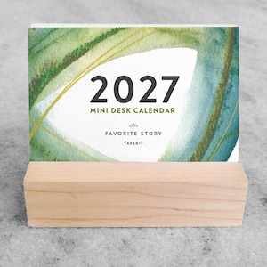 May include: A 2027 mini desk calendar featuring a watercolor design in shades of green and blue. The calendar is held in a rectangular light-colored wooden stand. The text reads 2027 MINI DESK CALENDAR, FAVORITE STORY, and PAPERIE.