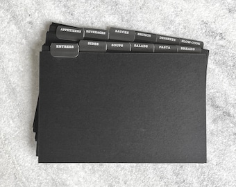 May include: A black recipe binder with dividers labeled with different categories of recipes, including appetizers, beverages, sides, soups, salads, sauces, brunch, desserts, pasta, breads, and slow cooker.