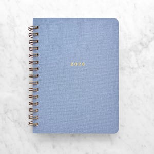 May include: A light blue spiral-bound notebook with the year 2026 in gold lettering. The notebook has a textured cover and a bronze-colored spiral binding. The notebook is on a white marble surface.
