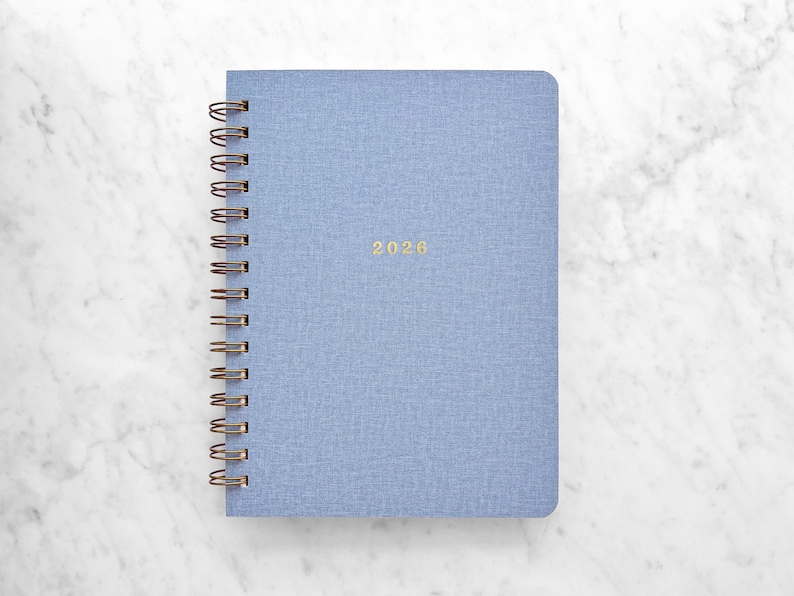 May include: A light blue spiral-bound notebook with the year 2026 in gold lettering. The notebook has a textured cover and a bronze-colored spiral binding. The notebook is on a white marble surface.