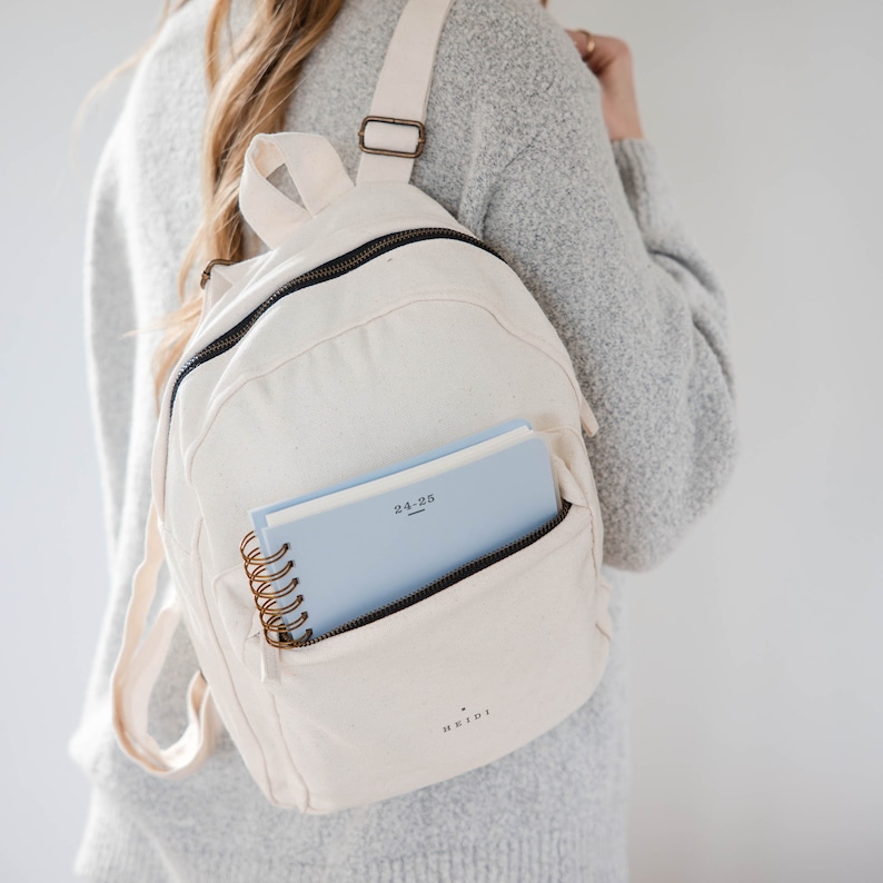 May include: A white canvas backpack with a zippered pocket and a blue notebook with the text "24-25" inside. The backpack has the word "Heidi" printed on the bottom.