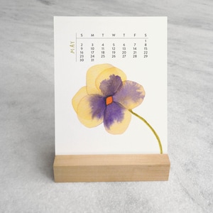 Peut inclure: Une carte calendrier rectangulaire blanche pour le mois de mai, pr&eacute;sentant une illustration &agrave; l'aquarelle d'une pens&eacute;e jaune et violette avec une tige verte. La carte est maintenue droite par un support en bloc de bois clair.