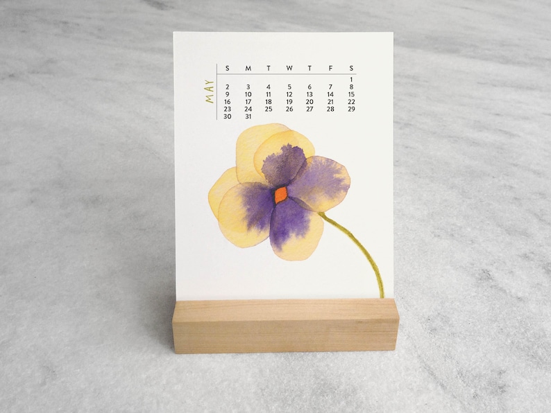 Peut inclure: Une carte calendrier rectangulaire blanche pour le mois de mai, pr&eacute;sentant une illustration &agrave; l'aquarelle d'une pens&eacute;e jaune et violette avec une tige verte. La carte est maintenue droite par un support en bloc de bois clair.