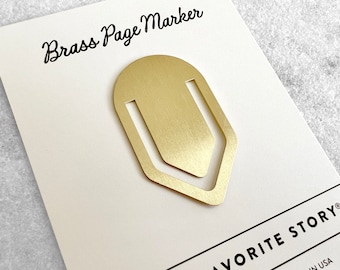 May include: A gold-colored brass page marker in the shape of a bookmark. The marker is on a white card with the text "Brass Page Marker" and "FAVORITE STORY" in black.