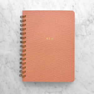 May include: A coral-colored spiral-bound notebook with gold-colored wire binding. The cover features the gold-colored initials "KSM". The notebook is on a white and gray marble surface.
