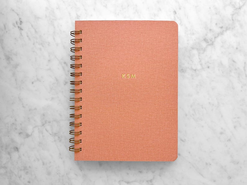 May include: A coral-colored spiral-bound notebook with gold-colored wire binding. The cover features the gold-colored initials "KSM". The notebook is on a white and gray marble surface.