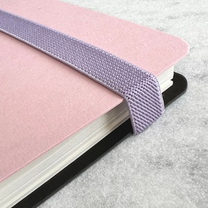 K&ouml;nnte beinhalten: Ein rosa Notizbuch mit einem lavendelfarbenen Gummibandverschluss. Das Notizbuch hat einen schwarzen R&uuml;cken und wei&szlig;e Papierseiten. Das Gummiband ist &uuml;ber das Notizbuch gespannt, um es geschlossen zu halten. Das Notizbuch liegt auf einer grauen Oberfl&auml;che.