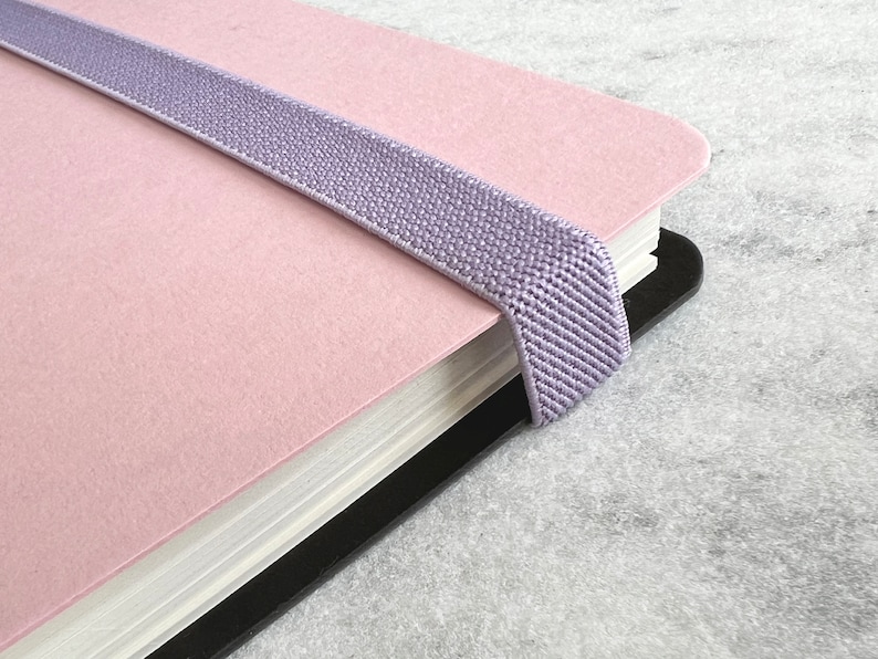 K&ouml;nnte beinhalten: Ein rosa Notizbuch mit einem lavendelfarbenen Gummibandverschluss. Das Notizbuch hat einen schwarzen R&uuml;cken und wei&szlig;e Papierseiten. Das Gummiband ist &uuml;ber das Notizbuch gespannt, um es geschlossen zu halten. Das Notizbuch liegt auf einer grauen Oberfl&auml;che.