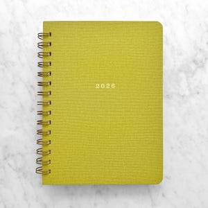 May include: A chartreuse-colored spiral-bound notebook with the year "2026" printed in gold. The notebook has a textured cover and gold-colored spiral binding. The notebook is on a white marble surface.
