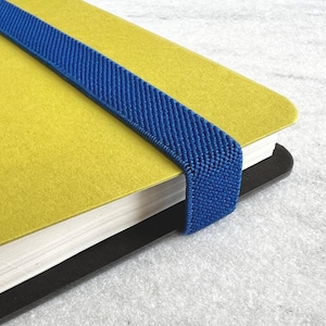 K&ouml;nnte beinhalten: Nahaufnahme eines gelben Notizbuchs mit einem blauen Gummiband. Das Notizbuch hat einen schwarzen R&uuml;cken und wei&szlig;e Seiten. Das Gummiband ist &uuml;ber den Einband gespannt.
