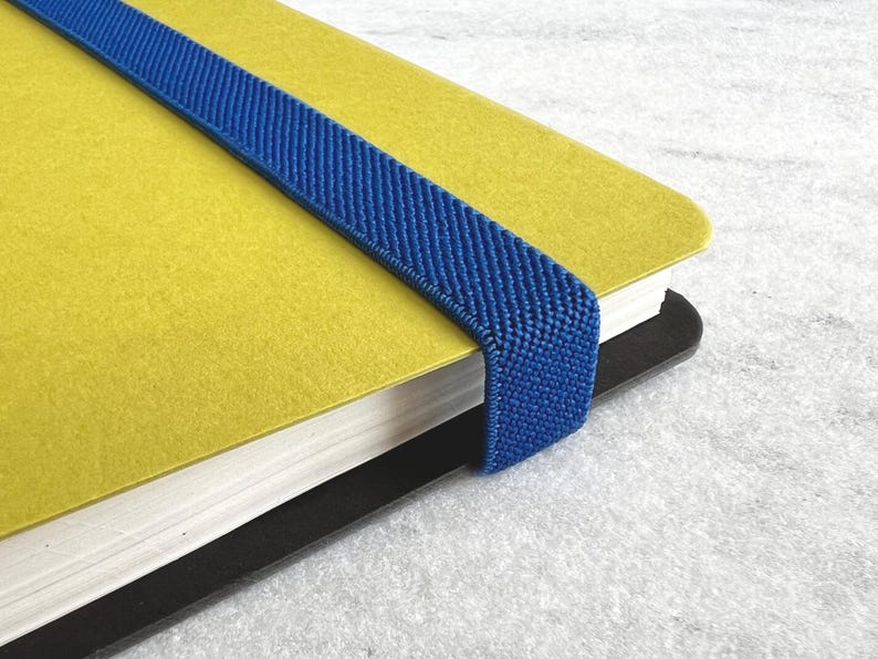 K&ouml;nnte beinhalten: Nahaufnahme eines gelben Notizbuchs mit einem blauen Gummiband. Das Notizbuch hat einen schwarzen R&uuml;cken und wei&szlig;e Seiten. Das Gummiband ist &uuml;ber den Einband gespannt.