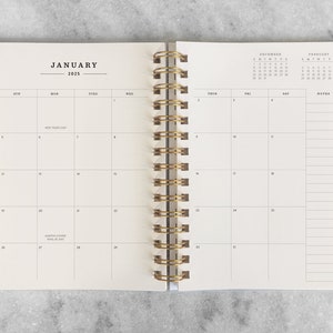 May include: A white planner with a gold spiral binding. The front cover has the word "JANUARY" and the year "2025" printed on it. The planner has a monthly calendar layout with space for notes.