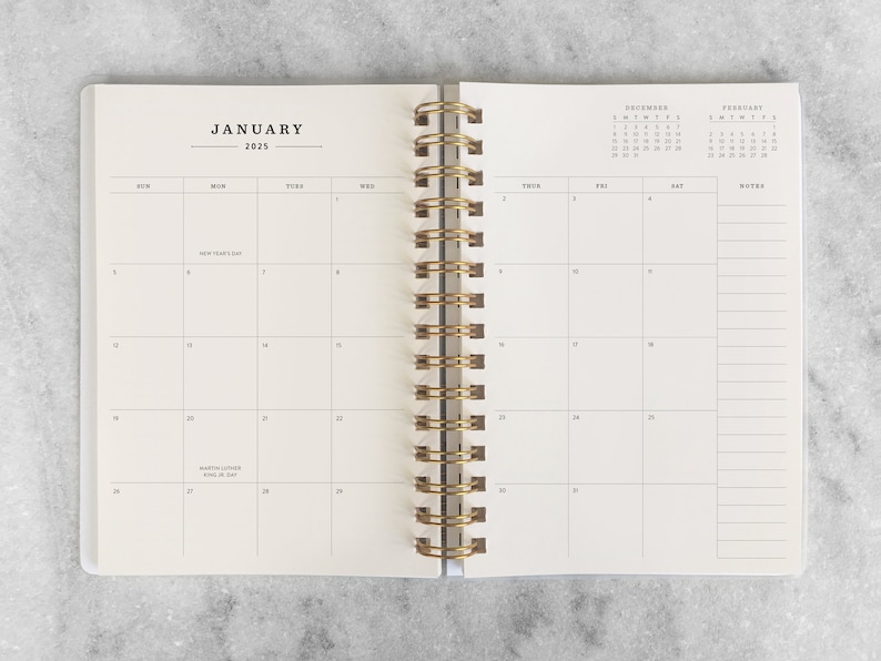 May include: A white planner with a gold spiral binding. The front cover has the word "JANUARY" and the year "2025" printed on it. The planner has a monthly calendar layout with space for notes.