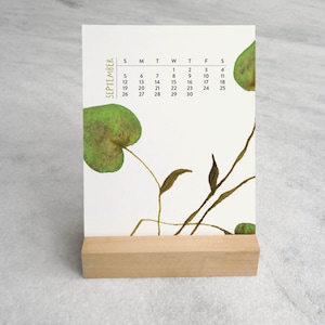 Peut inclure: Un calendrier de bureau vertical pour septembre avec un fond blanc, des illustrations de feuilles vertes et des tiges brunes fines. La grille du calendrier affiche les jours de la semaine et les dates en noir, le tout sur un socle en bois clair.