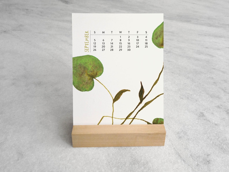 Peut inclure: Un calendrier de bureau vertical pour septembre avec un fond blanc, des illustrations de feuilles vertes et des tiges brunes fines. La grille du calendrier affiche les jours de la semaine et les dates en noir, le tout sur un socle en bois clair.