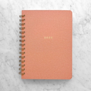 May include: A coral-colored spiral-bound notebook with the year "2026" in gold lettering. The notebook is set against a white and gray marble background. The spiral binding is a bronze color.