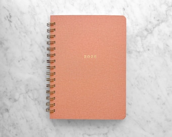 May include: A coral-colored spiral-bound notebook with the year "2026" in gold lettering. The notebook is set against a white and gray marble background. The spiral binding is a bronze color.