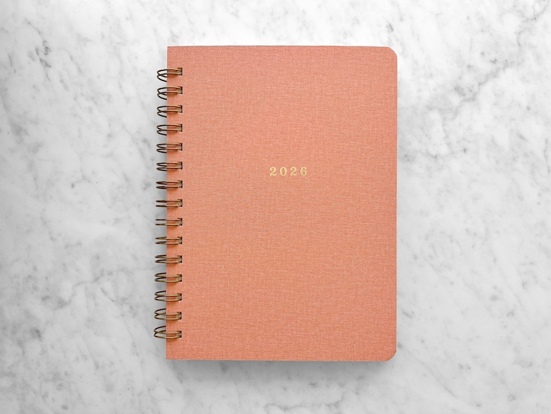 May include: A coral-colored spiral-bound notebook with the year "2026" in gold lettering. The notebook is set against a white and gray marble background. The spiral binding is a bronze color.