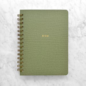 May include: A green spiral-bound notebook with gold-colored spiral binding and the initials "KSM" in gold lettering. The notebook has a textured cover and is set against a white marble background.