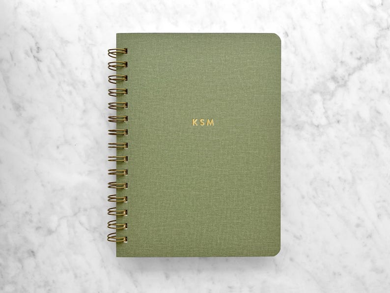 May include: A green spiral-bound notebook with gold-colored spiral binding and the initials "KSM" in gold lettering. The notebook has a textured cover and is set against a white marble background.