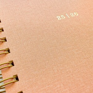 May include: Close-up of a spiral-bound notebook with a textured, peach-colored cover. The cover features the gold-colored text "25 | 26". The spiral binding is made of gold-colored metal.