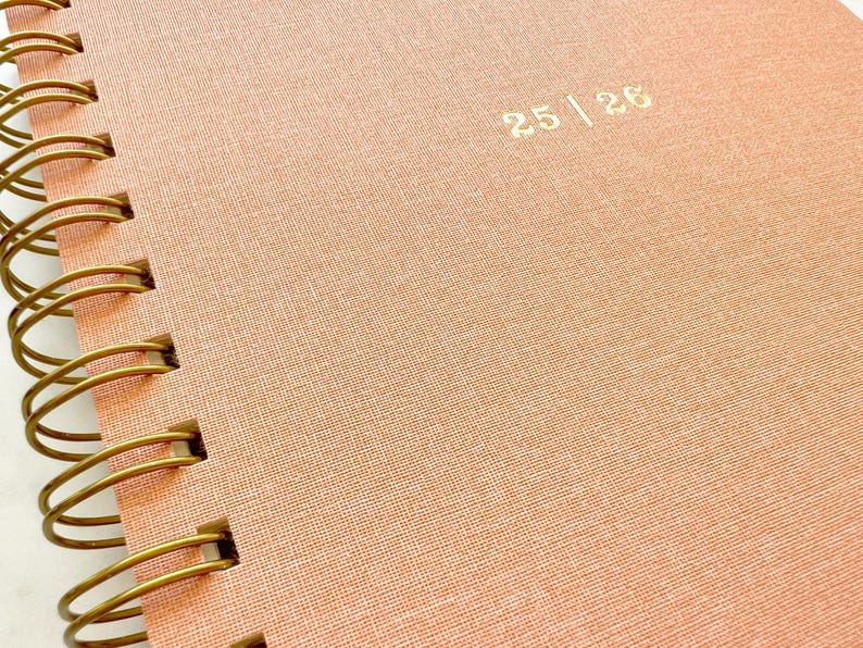 May include: Close-up of a spiral-bound notebook with a textured, peach-colored cover. The cover features the gold-colored text "25 | 26". The spiral binding is made of gold-colored metal.