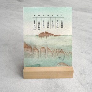May include: A vertical desk calendar for the month of May featuring a watercolor landscape in shades of seafoam green and brown. The calendar displays the days of the week and dates in black text, with the word May written in a cursive script.