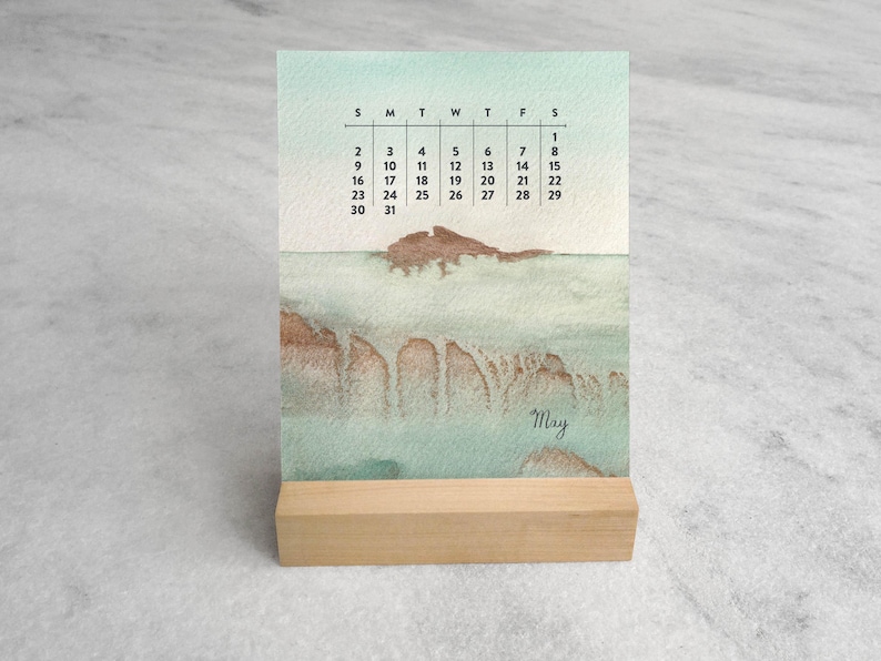 May include: A vertical desk calendar for the month of May featuring a watercolor landscape in shades of seafoam green and brown. The calendar displays the days of the week and dates in black text, with the word May written in a cursive script.