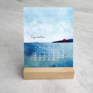 May include: A desk calendar for September featuring a watercolor painting of a blue sea and sky with a small red house on a dark cliff. The calendar grid is printed in white text over the water, and the card rests in a light wood block stand.