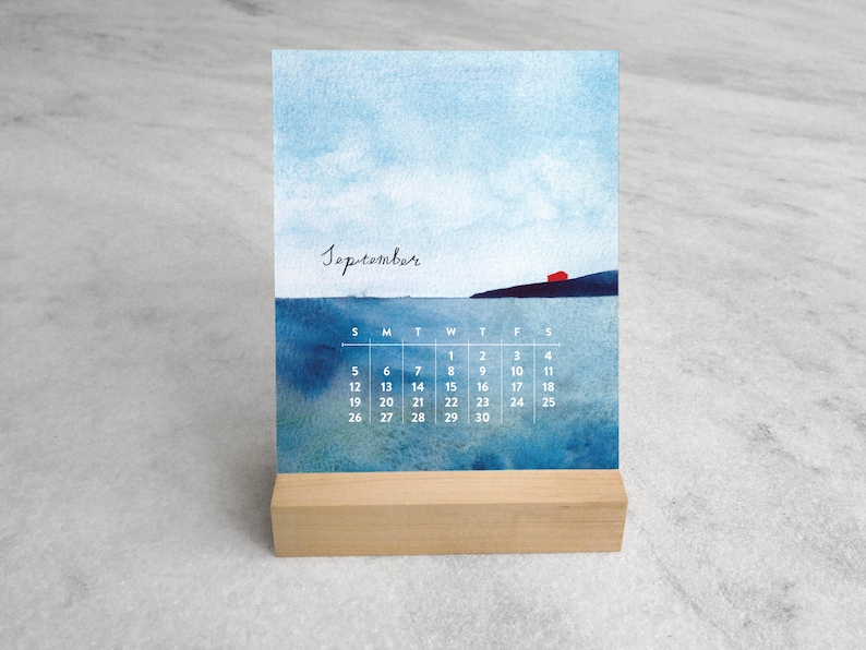 May include: A desk calendar for September featuring a watercolor painting of a blue sea and sky with a small red house on a dark cliff. The calendar grid is printed in white text over the water, and the card rests in a light wood block stand.