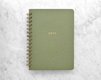 May include: A green spiral-bound planner with the year "2026" in gold lettering. The cover has a textured appearance. The planner is set against a white marble background.