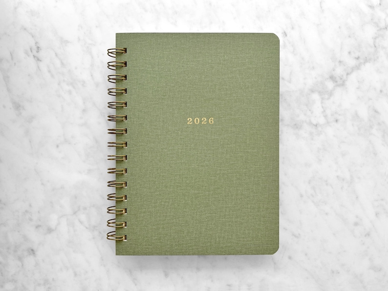 May include: A green spiral-bound planner with the year "2026" in gold lettering. The cover has a textured appearance. The planner is set against a white marble background.