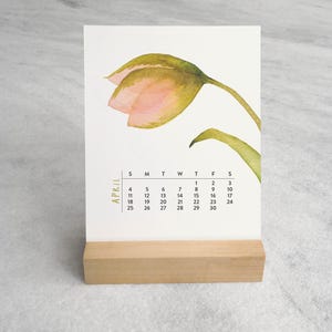 Peut inclure: Un calendrier de bureau avec une illustration aquarelle d'une tulipe rose et verte sur le dessus. Le mois d'avril est imprim&eacute; verticalement en lettres dor&eacute;es &agrave; c&ocirc;t&eacute; d'une grille noire indiquant les jours. La carte repose sur un socle en bois clair.