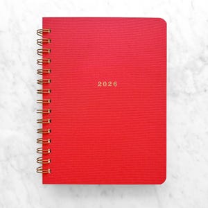 May include: A red spiral-bound notebook with gold-colored coils. The cover features the year "2026" in gold lettering. The notebook is set against a white marble background, suggesting a clean and organized aesthetic.