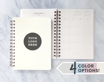 May include: A white spiral bound planner with a blank cover. The cover has a circle with the text "YOUR LOGO HERE". The planner has a calendar section with the months of December, January, and February. The planner is available in four color options.