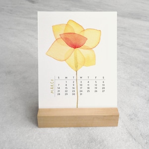 Peut inclure: Une carte calendrier rectangulaire blanche pour le mois de mars pr&eacute;sentant une illustration &agrave; l'aquarelle d'une jonquille jaune avec un centre orange. Le calendrier est pr&eacute;sent&eacute; sur un support en bois clair &agrave; base rectangulaire.