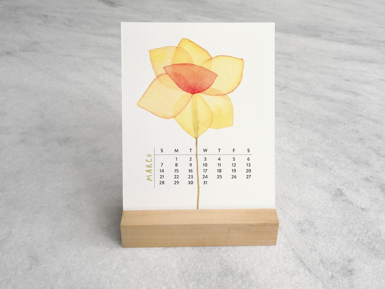 Peut inclure: Une carte calendrier rectangulaire blanche pour le mois de mars pr&eacute;sentant une illustration &agrave; l'aquarelle d'une jonquille jaune avec un centre orange. Le calendrier est pr&eacute;sent&eacute; sur un support en bois clair &agrave; base rectangulaire.