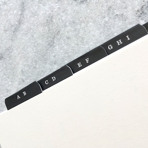 May include: A close-up of a notebook with black divider tabs labeled A through K. The tabs are arranged in a row, with each tab extending from the edge of the white paper. The letters are in a simple, sans-serif font.