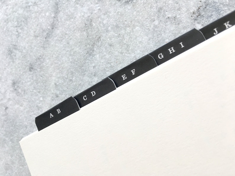 May include: A close-up of a notebook with black divider tabs labeled A through K. The tabs are arranged in a row, with each tab extending from the edge of the white paper. The letters are in a simple, sans-serif font.
