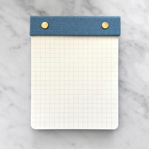 May include: A blue and white gridded notepad with brass fasteners. The notepad has a blue fabric cover.