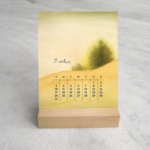 May include: A desk calendar featuring a watercolor illustration of a landscape with a green tree on a yellow hill. The text October is written in cursive above a grid displaying the days of the month, starting with Sunday through Saturday.