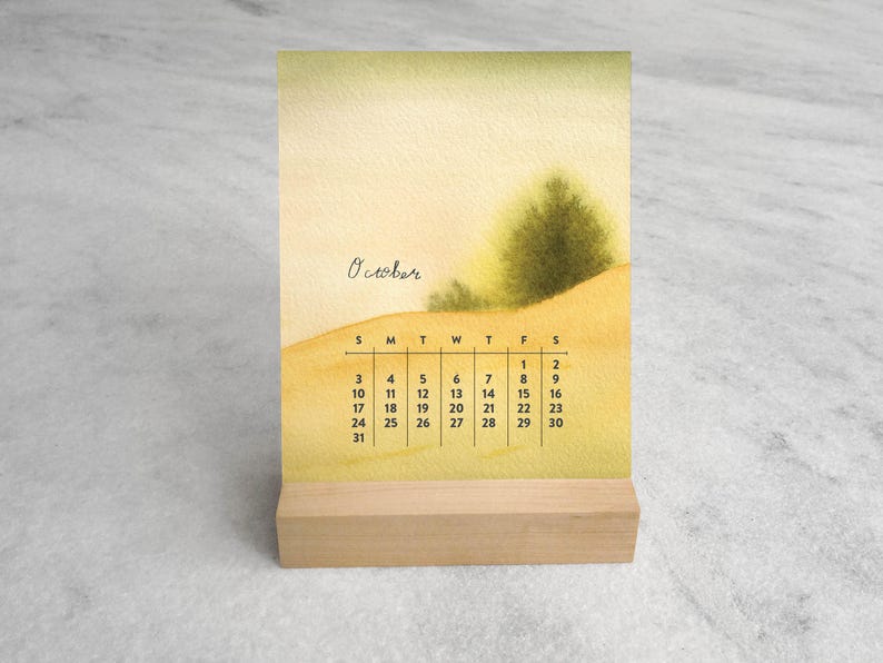 May include: A desk calendar featuring a watercolor illustration of a landscape with a green tree on a yellow hill. The text October is written in cursive above a grid displaying the days of the month, starting with Sunday through Saturday.