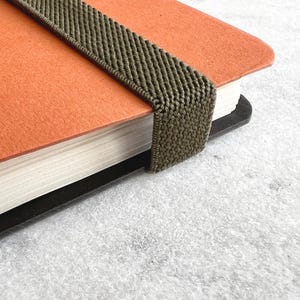 K&ouml;nnte beinhalten: Nahaufnahme eines orangefarbenen Notizbuchs mit einem dunkelgr&uuml;nen Gummibandverschluss. Die Seiten des Notizbuchs sind wei&szlig;, und der Einband hat eine einfarbige orange Farbe. Das Gummiband ist ein strukturiertes, gewebtes Material.