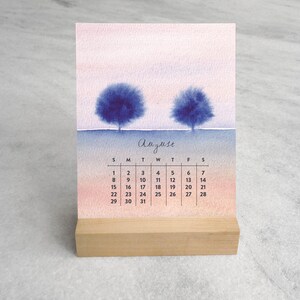 May include: A desk calendar for August featuring a watercolor illustration of two dark blue trees against a soft pink and blue gradient sky. The calendar grid is printed in black ink below the trees, and the entire card rests in a light wood stand.