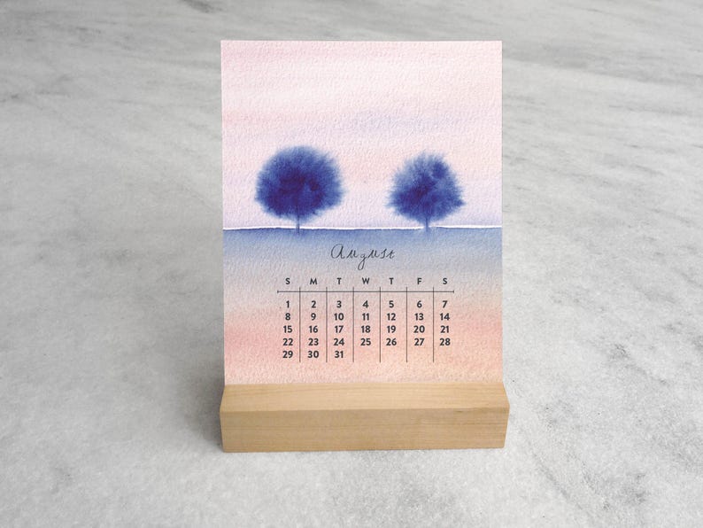 May include: A desk calendar for August featuring a watercolor illustration of two dark blue trees against a soft pink and blue gradient sky. The calendar grid is printed in black ink below the trees, and the entire card rests in a light wood stand.